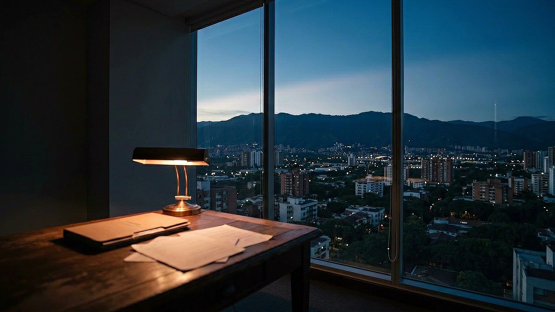 Escritorio de oficina con lampara encendida al atardecer, ventana con vista a Cali y las montanas al fondo